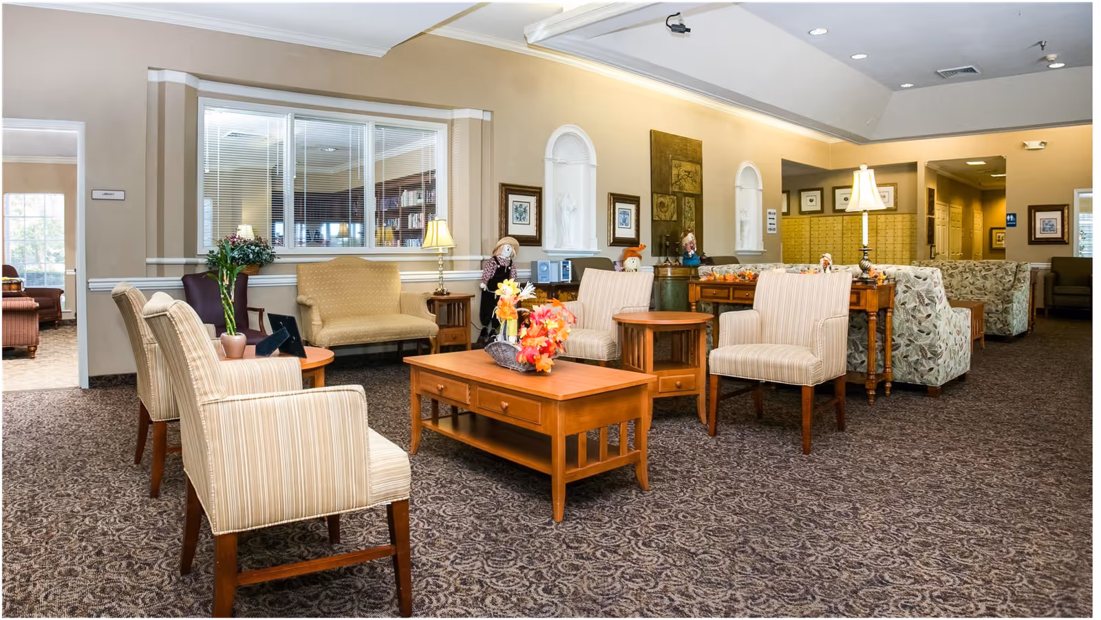 A spacious and well-lit common area with multiple seating options including striped armchairs, a beige loveseat, and floral-patterned sofas. There are wooden coffee tables with flower arrangements, table lamps, and decorative items. The walls are beige with framed artwork and small alcoves containing statues. A window looks into another room, and there is a hallway leading to other parts of the facility.