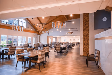 A spacious dining room with wooden floors, multiple tables and chairs arranged neatly, large windows letting in natural light, and modern chandeliers hanging from a high wooden ceiling.