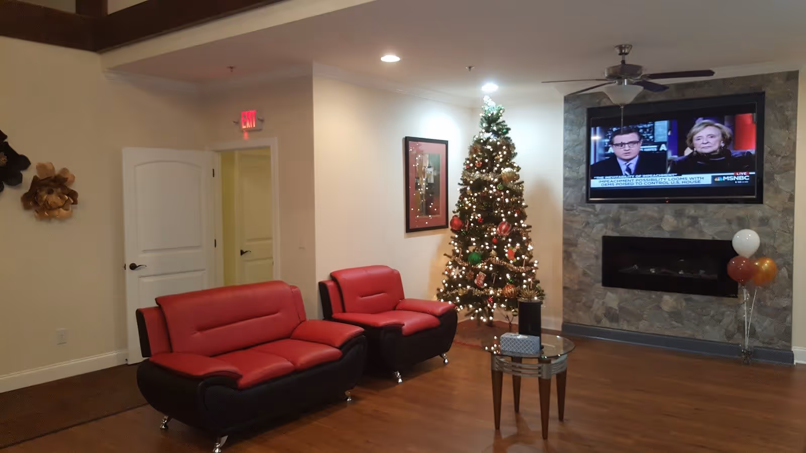 A cozy living room area with two red and black cushioned chairs, a decorated Christmas tree with lights and ornaments, a small glass coffee table with a tissue box, and a wall-mounted flat-screen TV showing a news program. The room has wooden flooring, a stone accent wall with a built-in fireplace, and a ceiling fan with a light fixture.