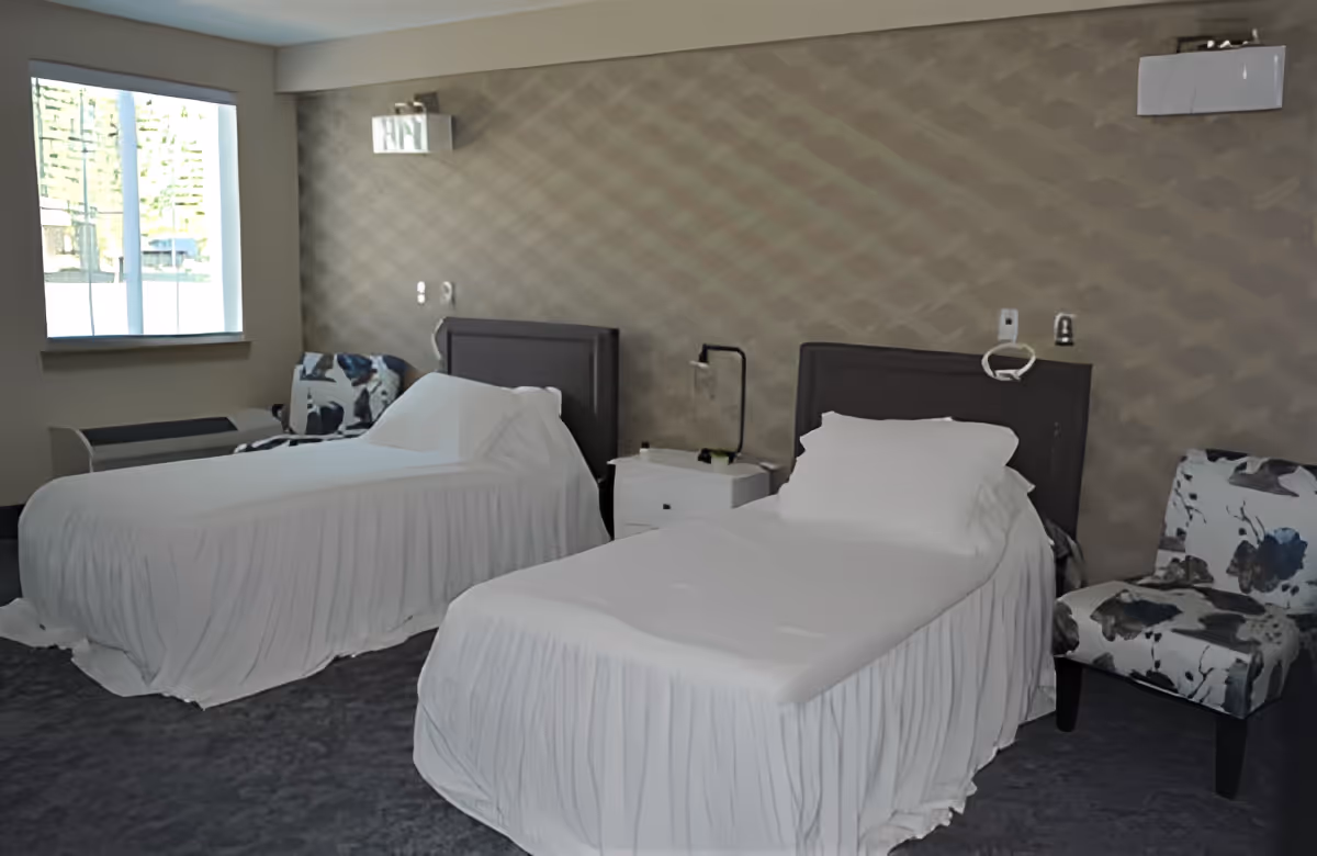 A room with two single beds covered in white sheets, each with a dark headboard. Between the beds is a white nightstand with a black lamp and some small items. There are two patterned chairs with floral designs, one near each bed. A window on the left side lets in natural light, and the walls have a textured, patterned wallpaper.