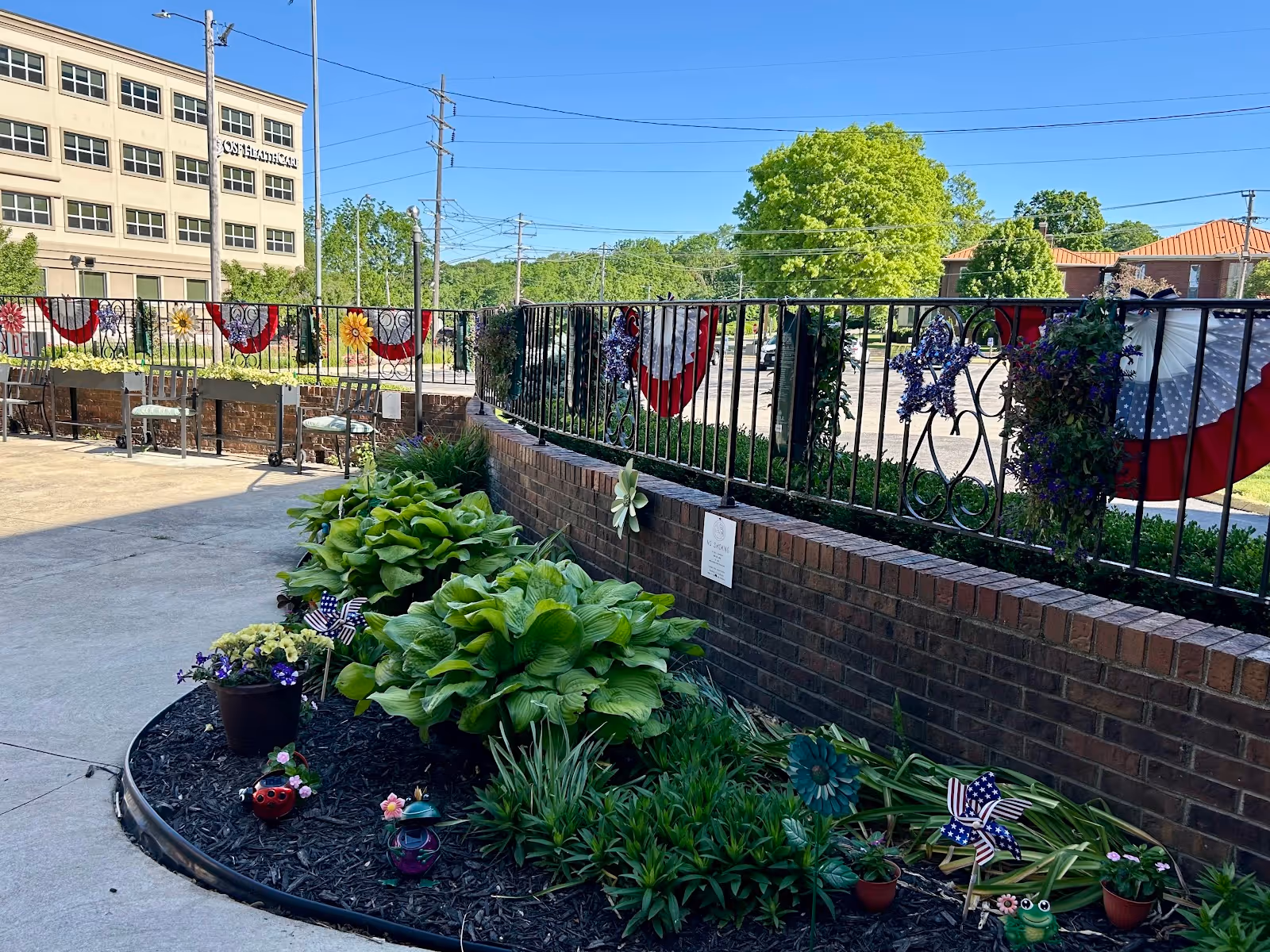 Outdoor patio area with a garden bed containing green plants and small decorative items like pinwheels and a ladybug ornament. The area is bordered by a brick wall with a black metal fence decorated with red, white, and blue bunting and star-shaped decorations. In the background, there is a parking lot, trees, and a multi-story building under a clear blue sky.