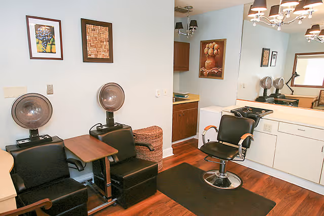 Interior of a hair salon area with two black salon chairs equipped with hair dryers, a small wooden table between them, a black styling chair in front of a large mirror, white cabinets, and framed artwork on the light blue walls.