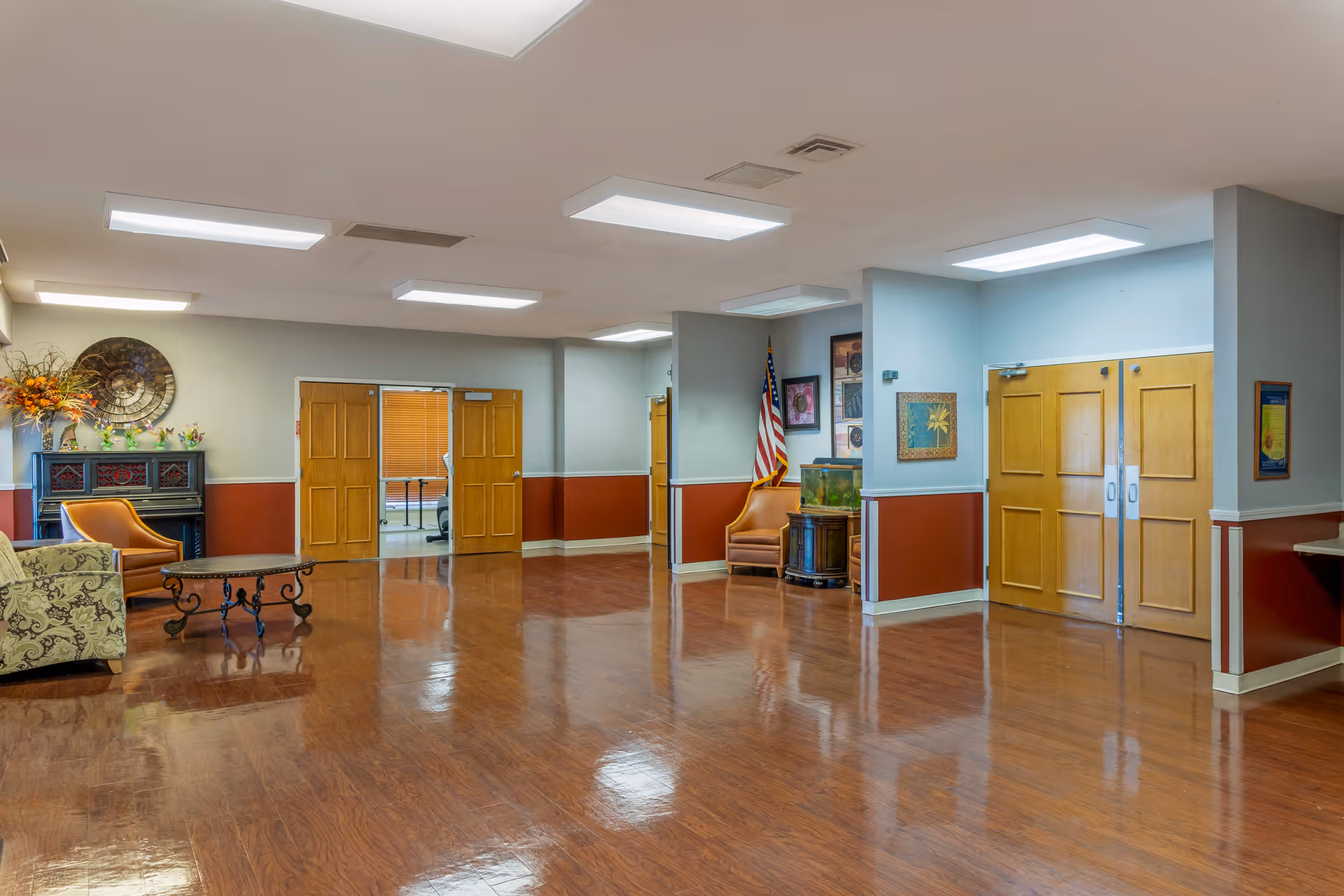 A spacious common area with polished wooden floors, beige and red walls, and multiple wooden double doors. The room is furnished with a patterned sofa, an orange armchair, a black coffee table, and a small table with a fish tank. There is an American flag in the corner and decorative artwork on the walls. The ceiling has several rectangular fluorescent lights.