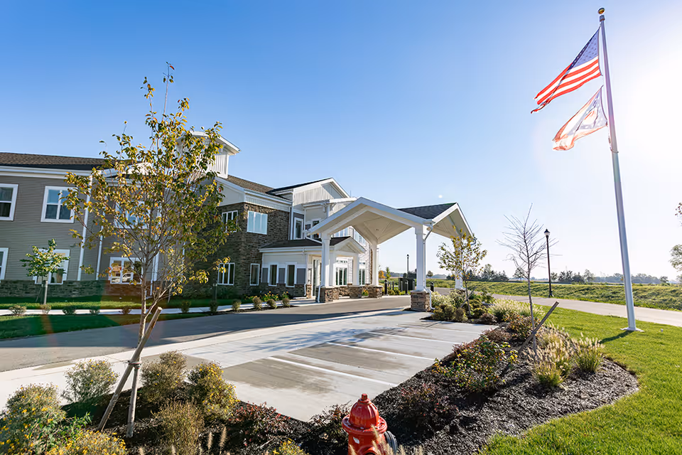Exterior view of The Oakwood Assisted Living at Otterbein Pemberville building on a clear sunny day, showing the entrance with a covered drop-off area, landscaped garden beds, a fire hydrant, and two flagpoles with the American flag and another flag flying.