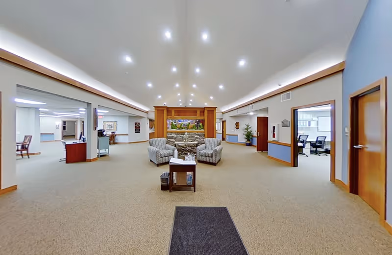 A spacious senior living facility common area with beige carpet and blue and beige walls. The room features two patterned armchairs and a small table with a water pitcher and glasses in the center. There is a decorative stone fireplace with a colorful painting above it. Several doorways lead to other rooms, including an office area with chairs and desks visible through one open door. The ceiling is white with recessed lighting.