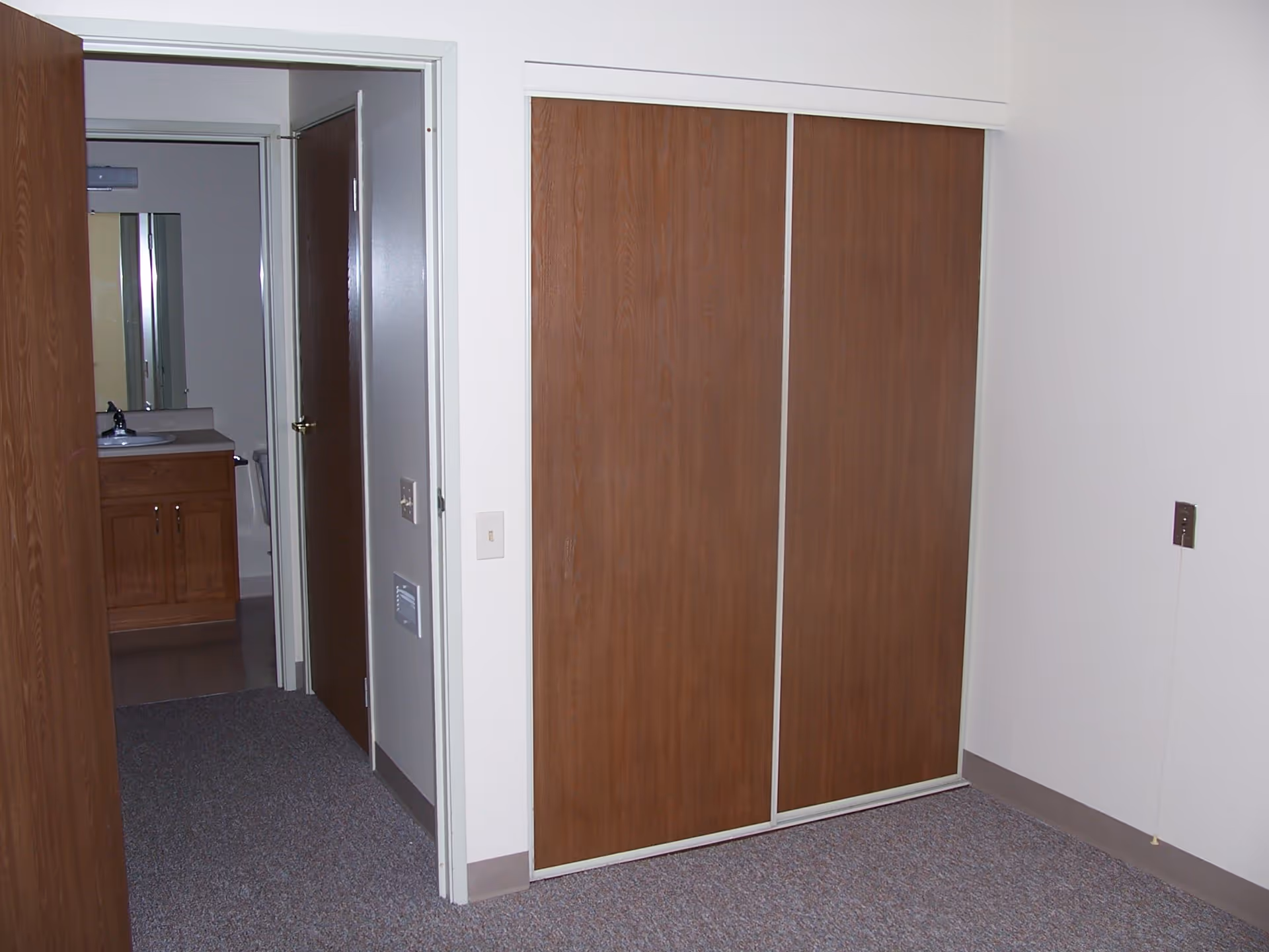 Interior view of a room with a carpeted floor, white walls, and a large wooden sliding door closet. There is an open doorway leading to a bathroom with a wooden vanity, sink, and mirror visible.
