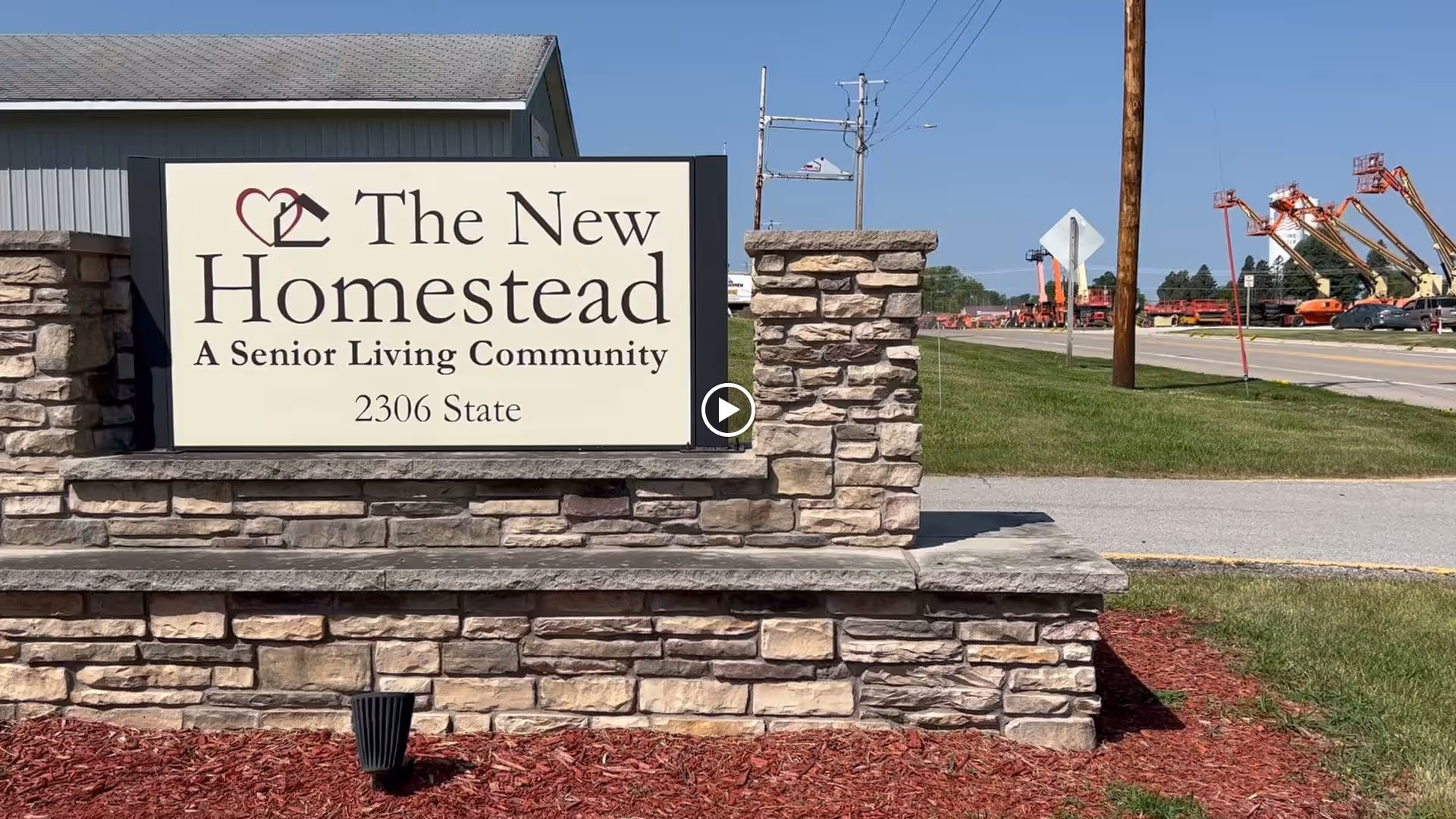 Stone roadside sign that reads "The New Homestead A Senior Living Community 2306 State" in front of a building and street.