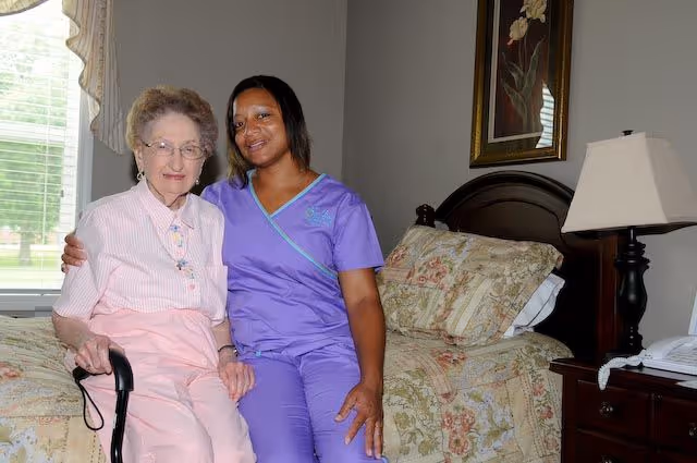An elderly woman sitting on the edge of a bed holding a cane, accompanied by a smiling caregiver in purple scrubs sitting beside her in a bedroom with floral bedding, a bedside table with a lamp and a telephone, and a window with curtains.