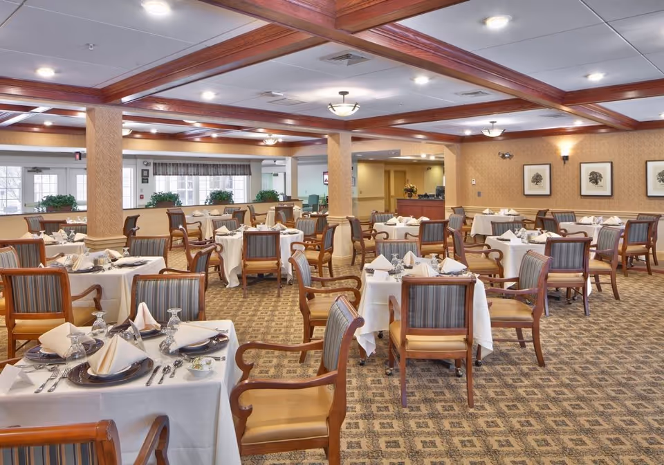 A spacious dining room with multiple tables set for a meal. Each table is covered with a white tablecloth and neatly arranged with plates, silverware, glasses, and folded napkins. The room features wooden beams on the ceiling, patterned carpet, and framed artwork on the walls. Large windows allow natural light to brighten the space.