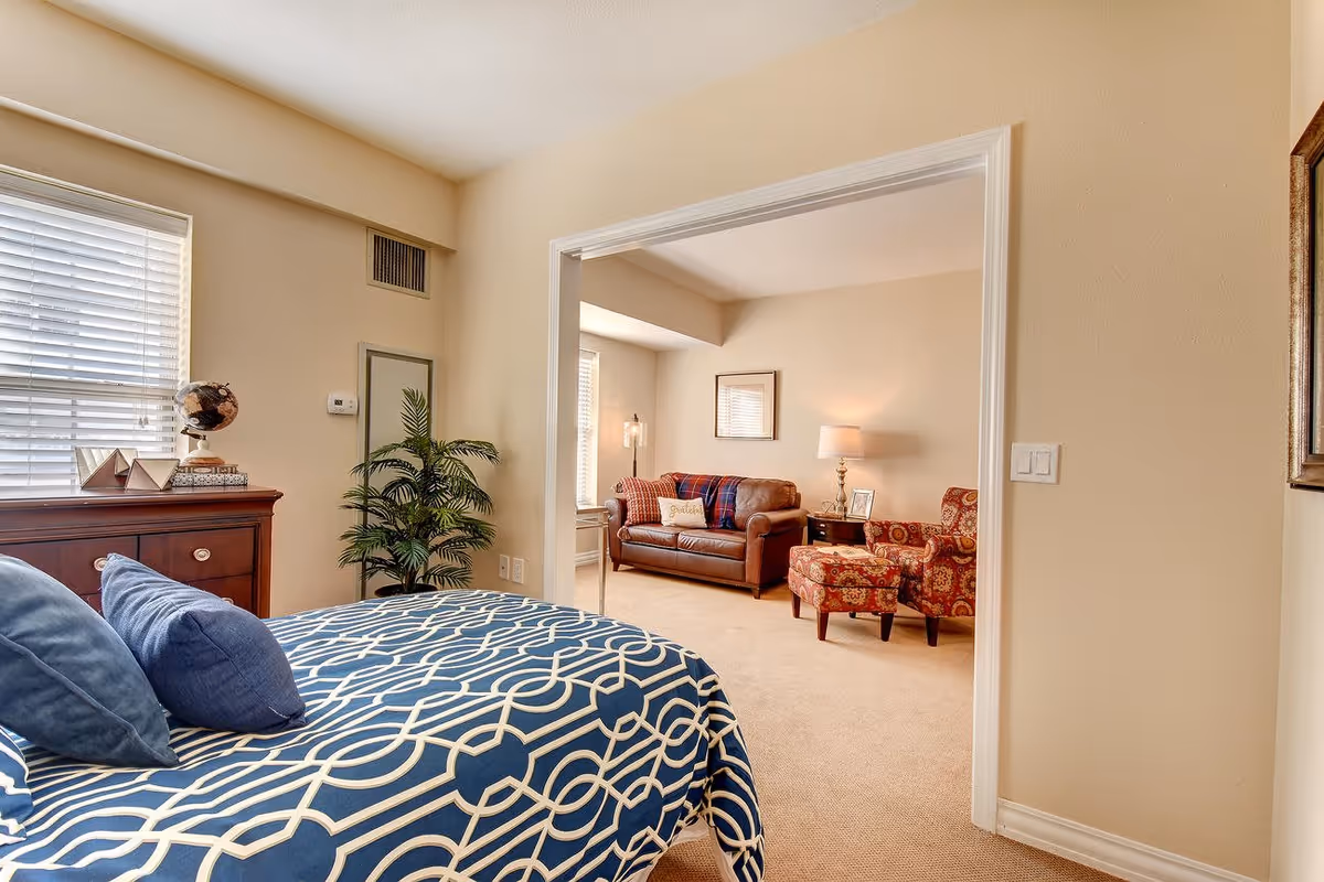 View from a bedroom into an adjacent living room area in a senior living facility. The bedroom features a bed with a blue and white patterned comforter and blue pillows, a wooden dresser with decorative items including a globe, and a potted plant. The living room has a brown leather sofa with a plaid throw and a pillow, a patterned armchair with a matching ottoman, a side table with a lamp and framed photo, and beige walls and carpet throughout.