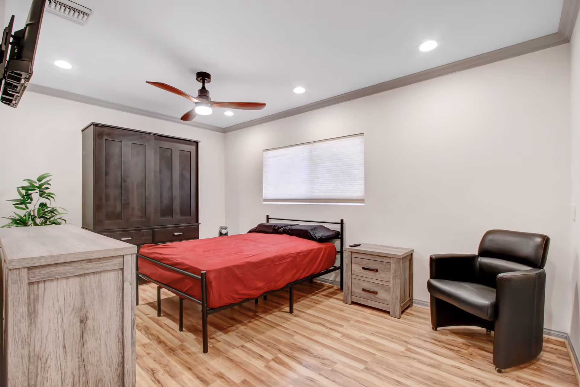 A clean and modern bedroom with a wooden floor, a bed with a red bedspread and black pillows, a wooden dresser, a dark wooden wardrobe, a black leather armchair, a small wooden nightstand, a ceiling fan with wooden blades, and a window with a white blind.