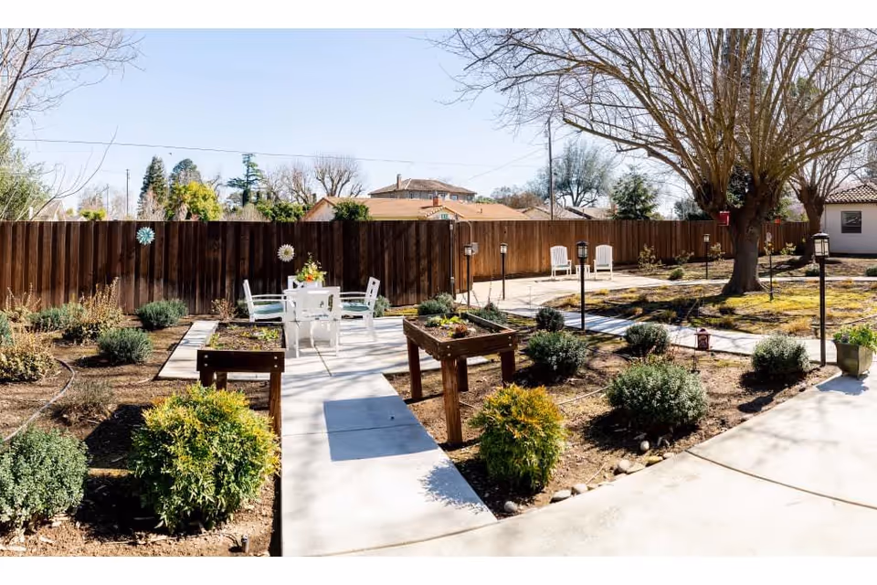 Outdoor garden area with raised planting beds, shrubs, and a paved walkway. There is a white table with four chairs in the center, and two white chairs near a large tree. A wooden fence encloses the space, and there are several small lantern-style lights along the walkway.