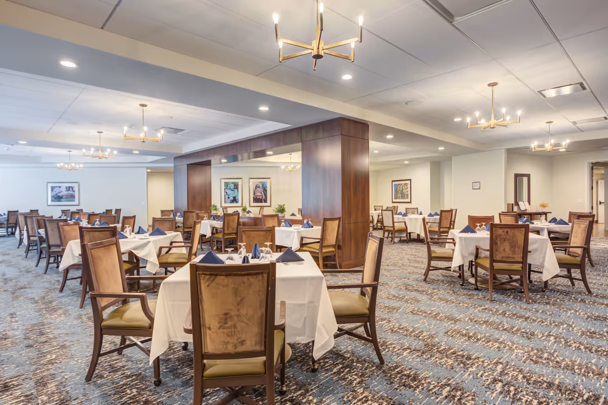 A spacious dining room with multiple tables covered in white tablecloths and set with blue napkins and glassware. The room features wooden chairs with cushioned seats, modern chandeliers hanging from the ceiling, and framed artwork on the walls. The carpet has a patterned design in shades of blue and brown.