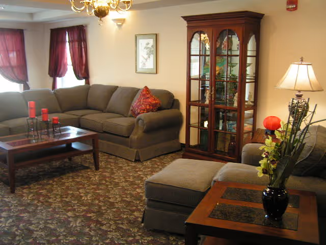 A cozy living room with a large gray sectional sofa adorned with a red decorative pillow, a wooden coffee table with red candles, a glass-front wooden display cabinet, a side table with a vase of flowers, and a lamp. The room has patterned carpet, beige walls, and windows with red curtains.