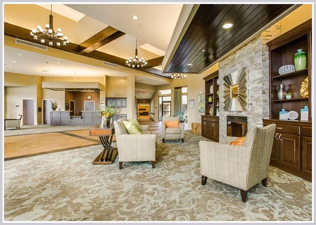 A spacious and well-lit common area in a senior living facility featuring comfortable armchairs with cushions arranged around a small wooden table. The room has a patterned carpet, a stone fireplace with decorative items on built-in wooden shelves, and modern chandeliers hanging from a ceiling with wooden beams. In the background, there is a reception desk and a hallway leading to other parts of the facility.