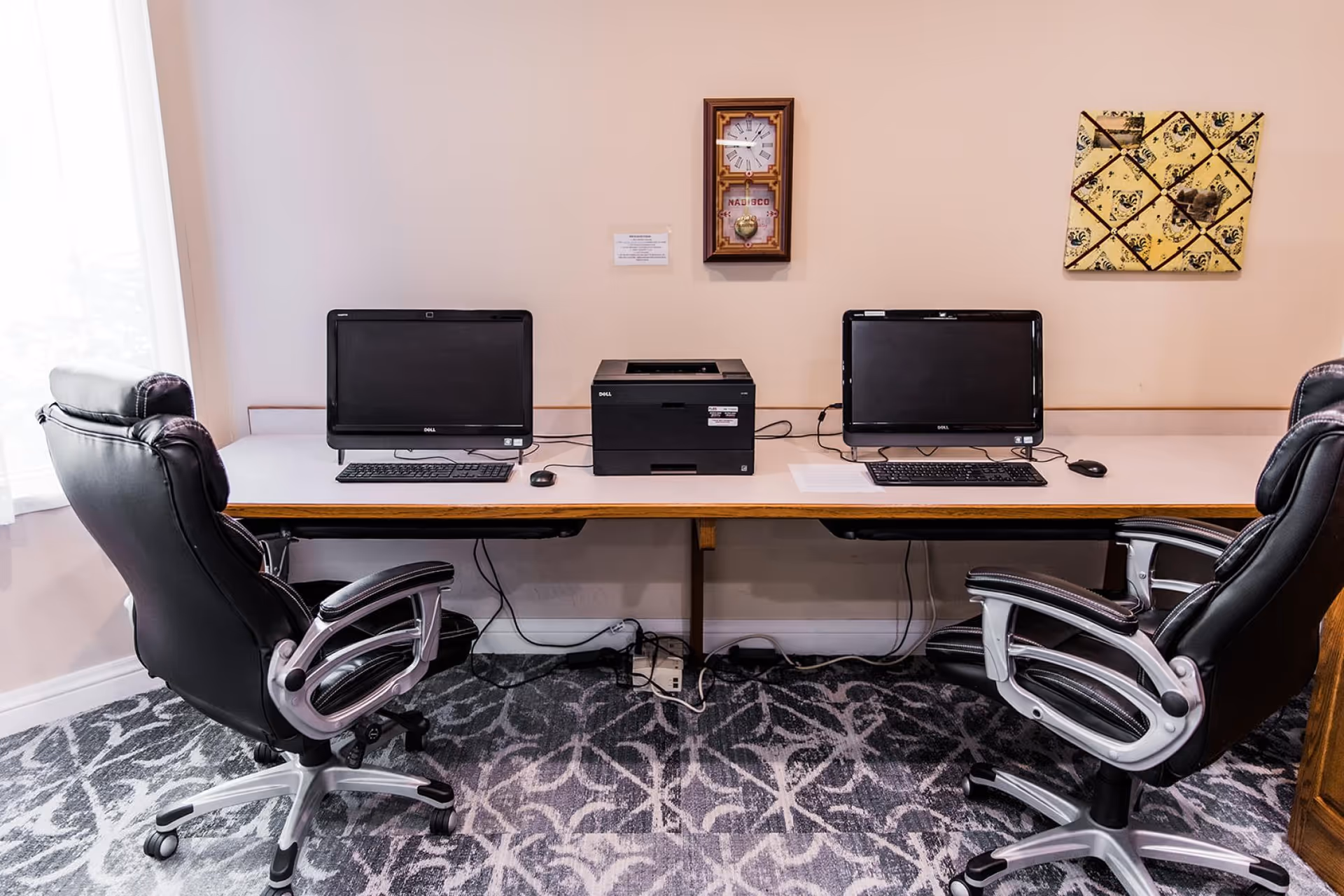 A small office or computer workstation area with two black leather office chairs, two Dell desktop computers, and a Dell printer placed on a long white desk against a beige wall. A window with white curtains is on the left side, and there is a decorative clock and a bulletin board on the wall above the desk. The floor is carpeted with a patterned design.