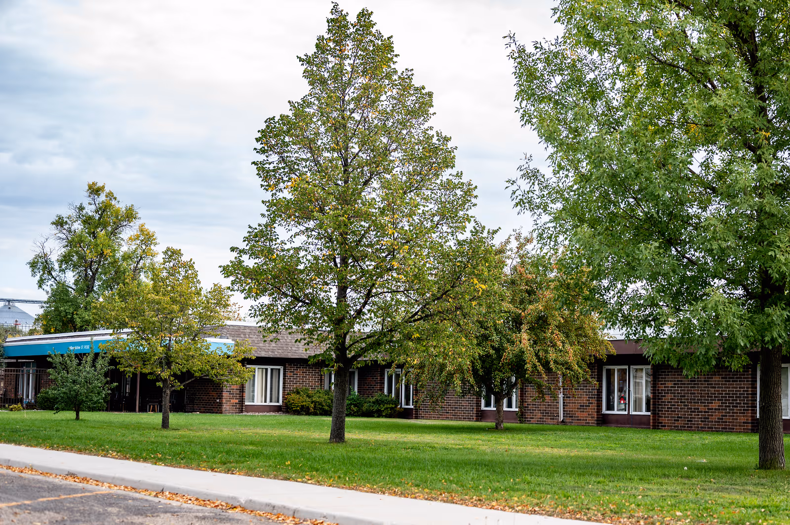 Exterior view of a single-story brick building with several windows, surrounded by green grass and multiple leafy trees under a cloudy sky.