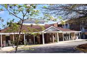 Front entrance of a brick senior living building with a covered drive, white columns, and rocking chairs on a porch.