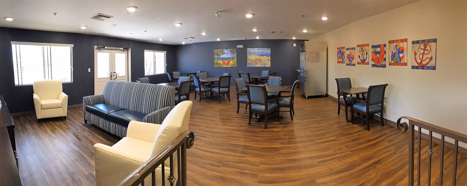 A spacious common area with wooden flooring, featuring multiple tables and chairs arranged for dining or socializing. There are two white armchairs and a striped sofa near the windows and door. The walls are decorated with colorful paintings and nautical-themed artwork. The room is well-lit with ceiling lights and natural light from the windows.