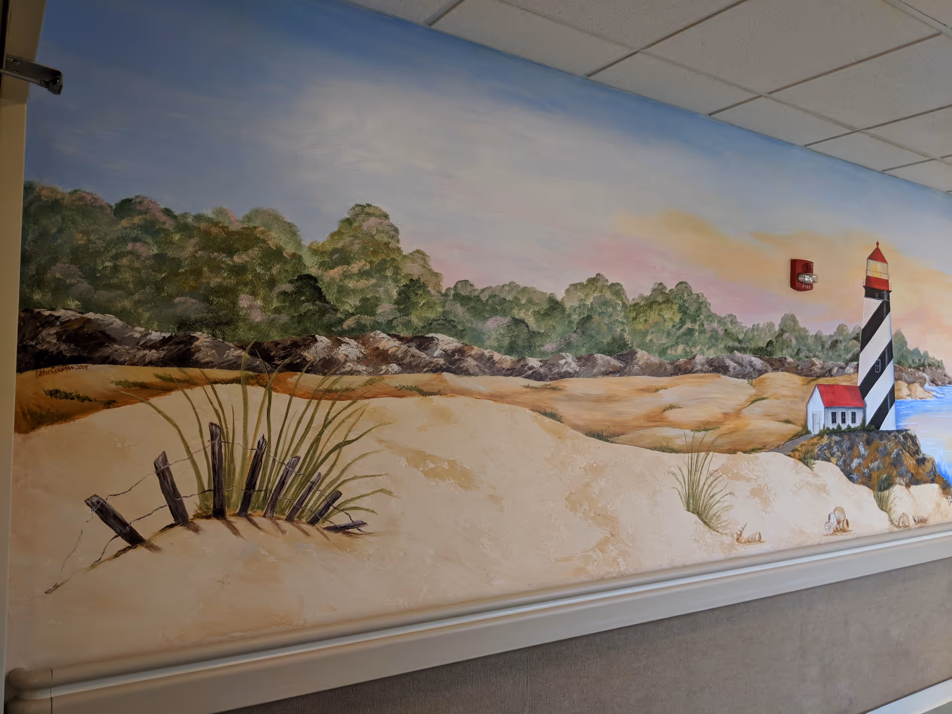 A hallway wall mural depicting sandy dunes, grasses, trees and a striped lighthouse under a pastel sky.
