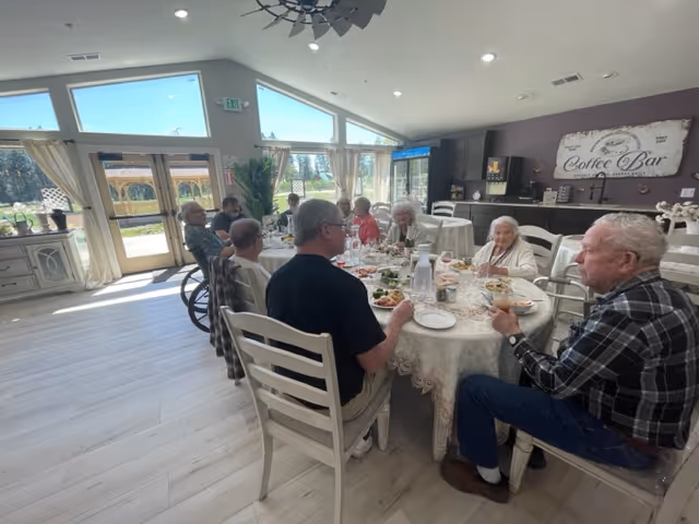A group of elderly people sitting around a round table enjoying a meal together in a bright room with large windows and a door leading outside. The room has light-colored wooden floors, a coffee bar area in the background, and a sign that reads 'Coffee Bar' on the wall.