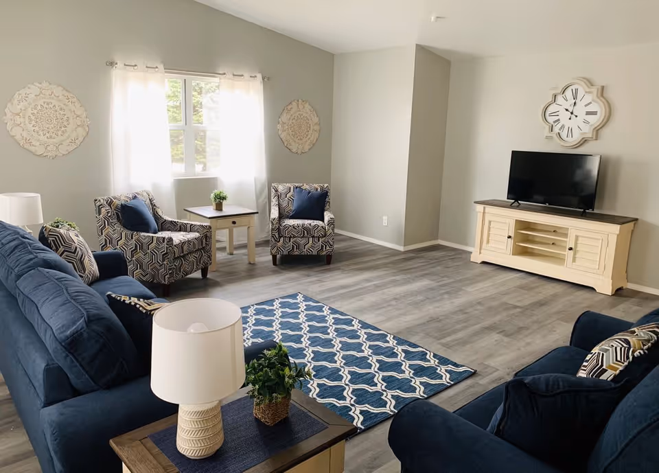 A bright living room with two blue sofas, two patterned armchairs, a wooden coffee table with a lamp and a small plant, a blue patterned rug, a TV on a cream-colored stand, and a decorative wall clock above the TV. The room has light gray walls and a window with sheer white curtains letting in natural light.