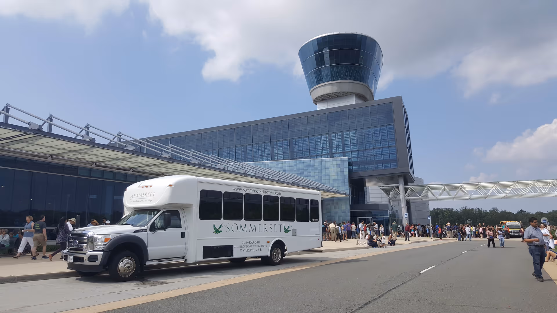 A white Sommerset Retirement Community shuttle bus parked outside a modern glass building with a unique circular tower on top. Many people are gathered on the sidewalk near the building entrance under a partly cloudy sky.