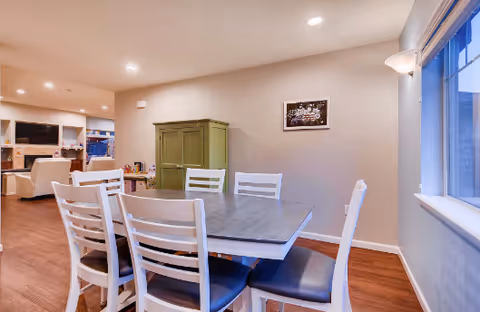 Interior view of a senior living facility dining area with a rectangular table surrounded by six white chairs with dark cushions. The room has wooden flooring, beige walls, a green cabinet against the wall, and a window on the right side. In the background, there is a living room area with a fireplace, TV, and additional seating.