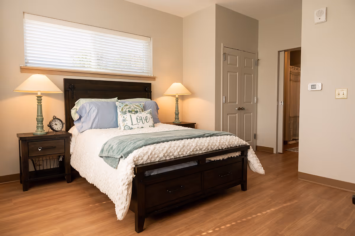 A cozy bedroom with a wooden bed frame and headboard, neatly made with white textured bedding, blue pillows, and a decorative pillow with the word 'Love'. There are two matching bedside tables with green lamps and an alarm clock on the left table. A window with white blinds is above the headboard. The room has wooden flooring, beige walls, and a door leading to a bathroom and a closet.