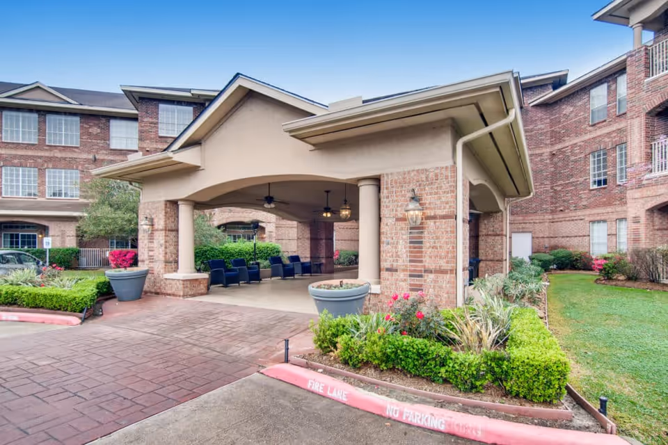 Entrance area of The Terraces at Kingwood Town Center senior living facility showing a covered drop-off zone with seating, surrounded by brick buildings and landscaped greenery under a clear blue sky.
