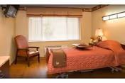 A cozy bedroom in a senior living facility featuring a single bed with a red quilted bedspread, a wooden chair with a cushion, a window with blinds, a bedside table with a lamp, and a wall-mounted television.