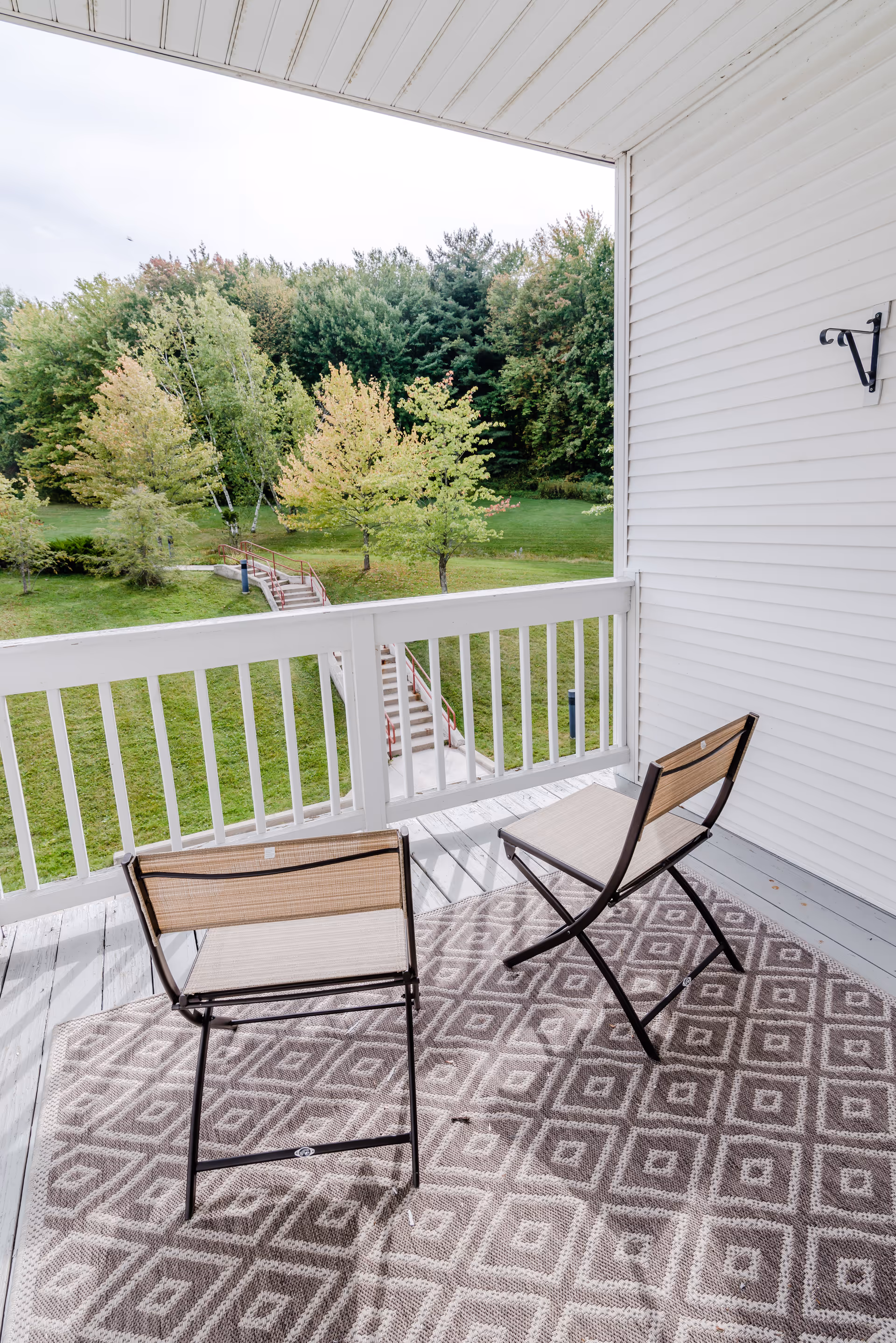 Two chairs on a patterned rug on a covered balcony overlooking a grassy yard and trees.