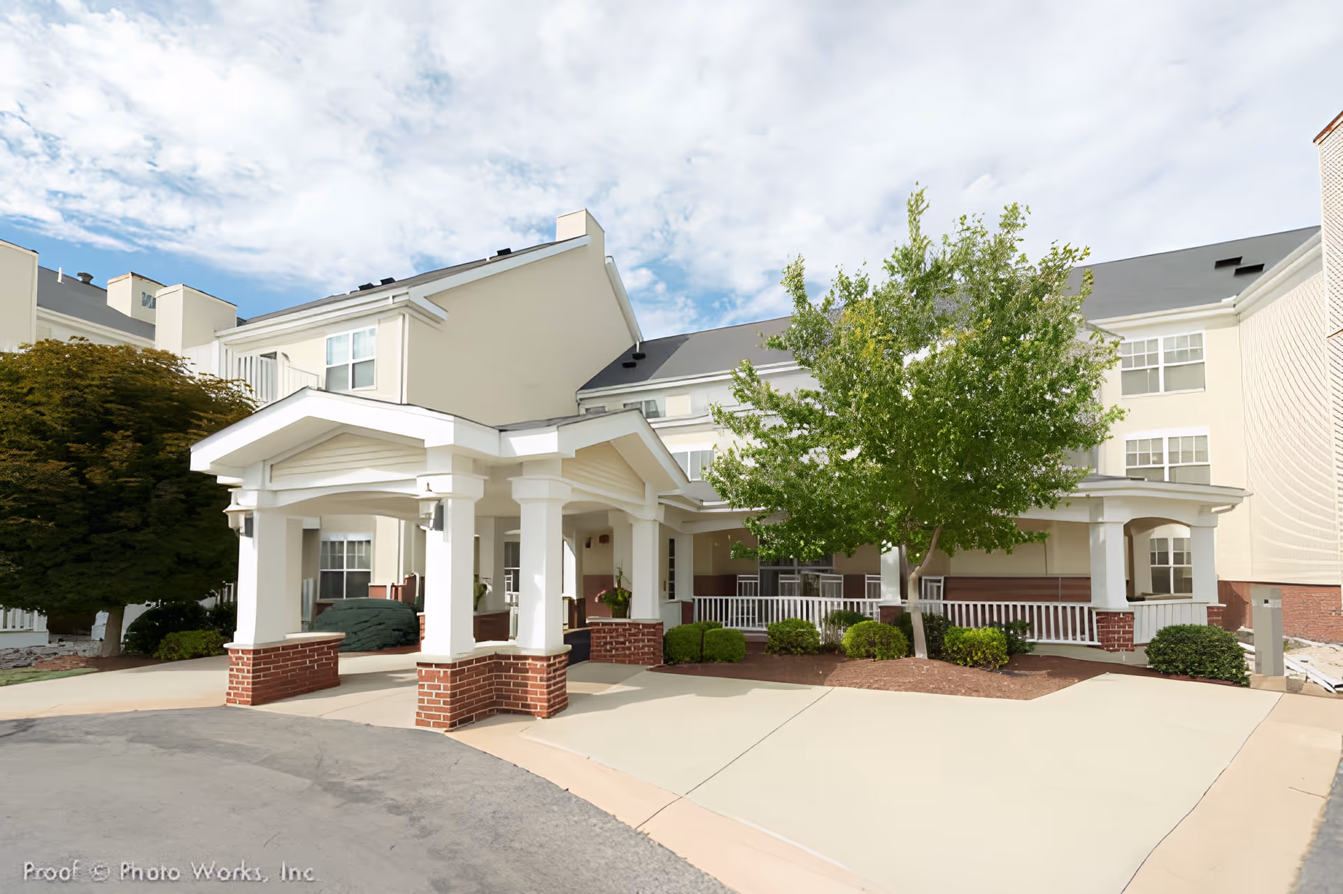 Exterior view of The Commons of Providence senior living facility in Sandusky, OH, showing a covered entrance with white columns and brick bases, surrounded by trees and shrubs under a partly cloudy sky.