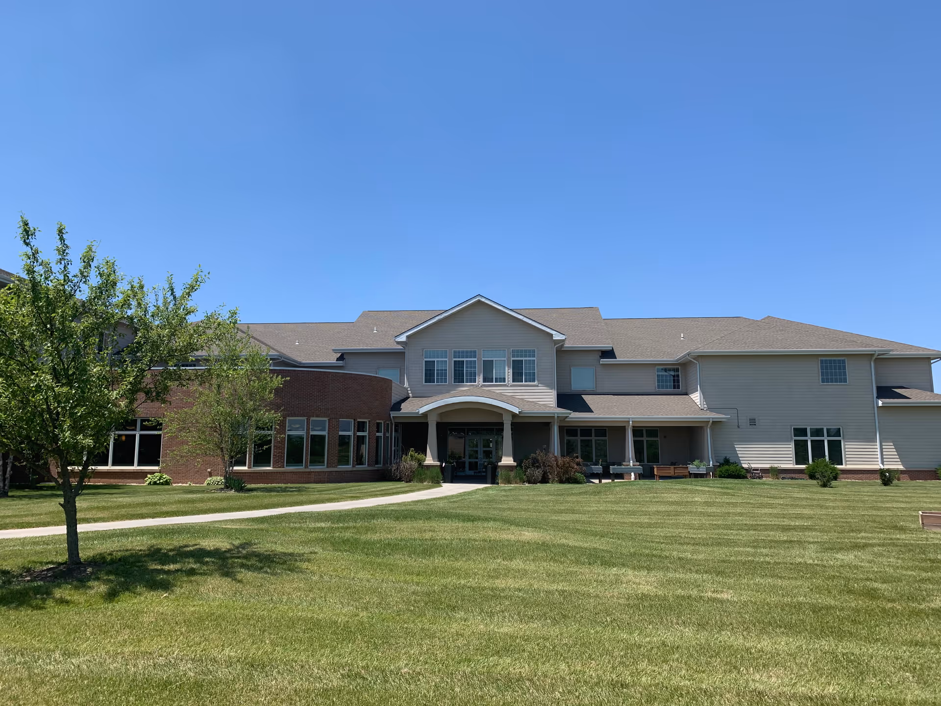 Exterior view of a large senior living facility building with a well-maintained lawn and a few trees under a clear blue sky.