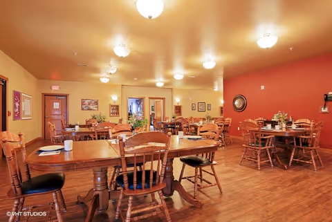 Warm dining room with multiple wooden tables and chairs, place settings, and soft ceiling lights.