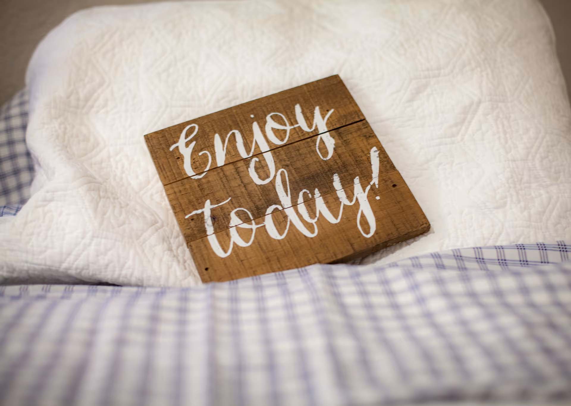 A wooden sign with white cursive text that reads 'Enjoy today!' resting on a white quilted blanket with a blue and white checkered fabric at the bottom edge.