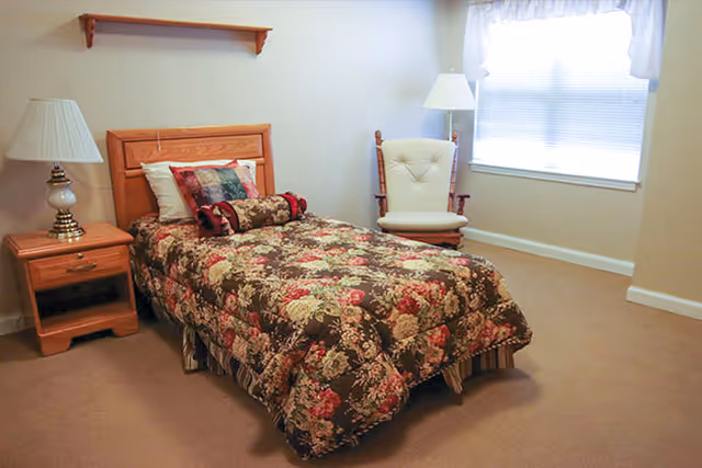 A bedroom with a single bed covered in a floral patterned comforter and multiple pillows. Next to the bed is a wooden nightstand with a lamp. There is a cushioned rocking chair and a floor lamp near a window with white curtains.