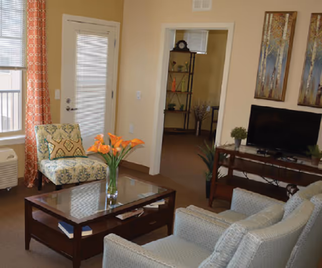 Cozy furnished living room with armchairs, a patterned accent chair, glass coffee table with orange flowers, TV on a stand, and a doorway to another room.