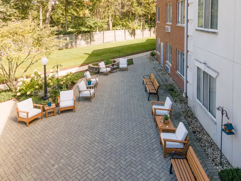 Outdoor patio area with cushioned wooden chairs, small tables with plants, and benches along the side of a brick and white building. The patio is paved with interlocking bricks and surrounded by greenery and trees.