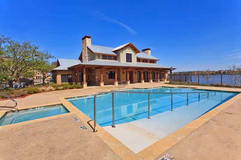 Outdoor swimming pool with a metal handrail and a smaller adjacent hot tub, set in front of a large building with a covered porch and stone chimneys under a clear blue sky.