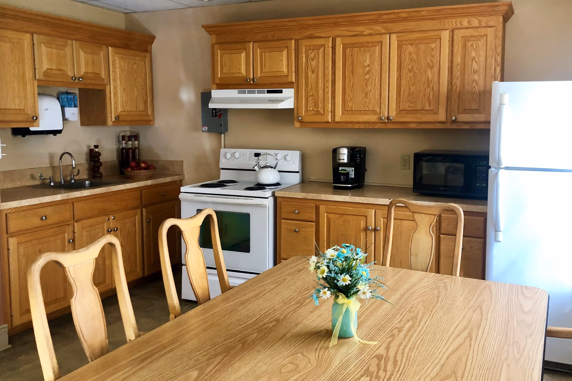 Wood-paneled kitchen with oak cabinets, a white stove and refrigerator, and a dining table with chairs and a small vase of flowers.