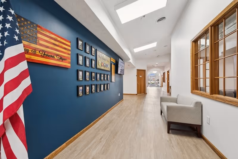 A bright hallway in Five Star Residences of Lafayette with light wood flooring and white walls. On the left side, a dark blue accent wall displays framed photos, plaques, and an American flag. On the right side, there is a beige upholstered bench beneath a large wooden-framed window. The hallway is illuminated by ceiling lights and leads to a door and a distant window.
