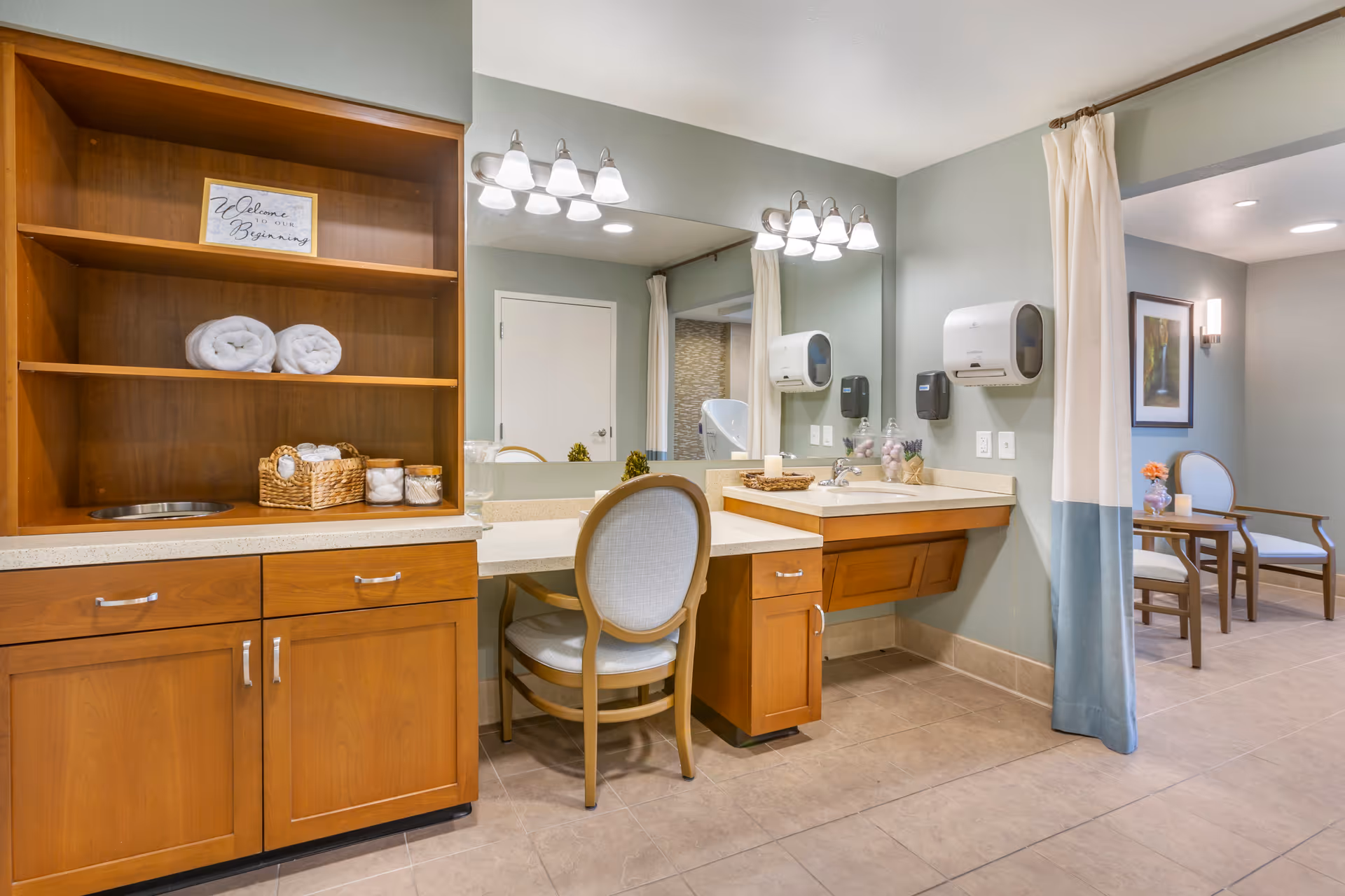 A well-lit interior space featuring a wooden vanity with a large mirror, two sets of light fixtures, and a cushioned chair. The vanity has a sink with soap dispensers mounted on the wall. To the left, there is a wooden shelving unit with rolled towels, a basket with cotton pads, and a small sign that reads 'Welcome to our Beginning.' A curtain partially separates this area from a seating area with two chairs and a small table with a flower vase and candle.