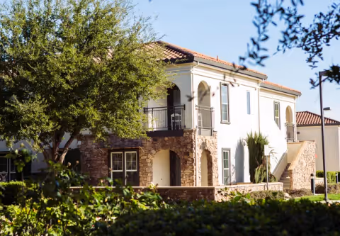 Two-story Mediterranean-style residential building with arched balconies, stone accents, and landscaped trees and shrubs.