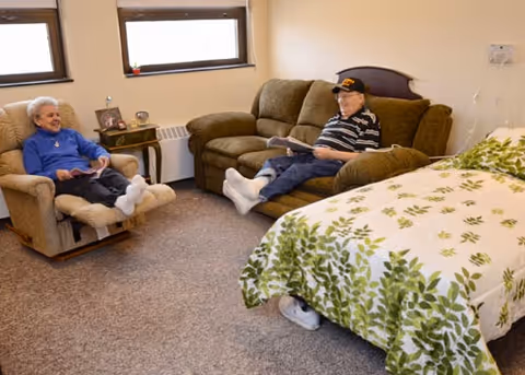 Two elderly people relaxing in a cozy room with a bed covered in a green and white leafy patterned blanket. One person is seated in a recliner chair, and the other is sitting on a brown couch. The room has beige walls, two windows, and a small table with picture frames and a clock.