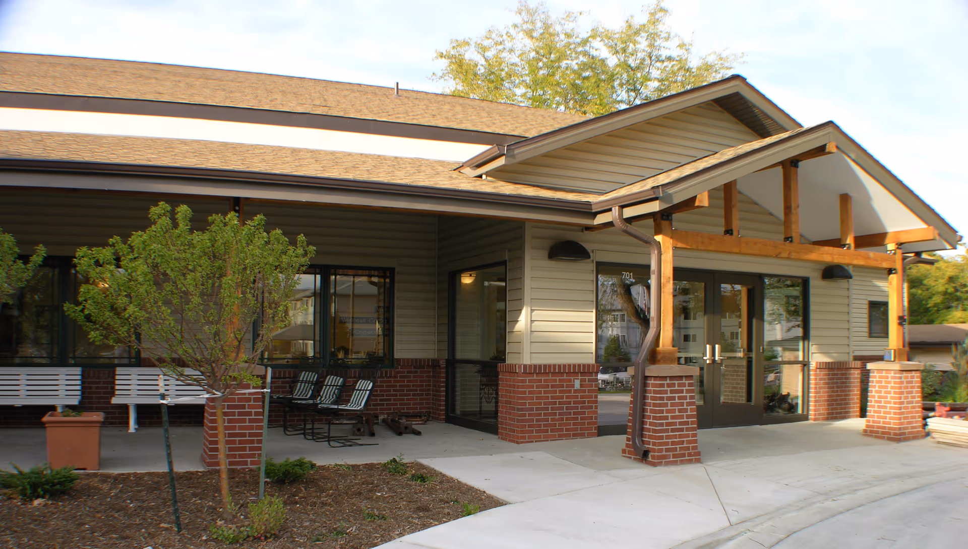 Exterior view of the entrance to Good Samaritan Society - Bonell Community, featuring a covered porch with brick pillars, glass double doors, and outdoor seating including white benches and striped chairs. There are small trees and landscaping in front of the building.
