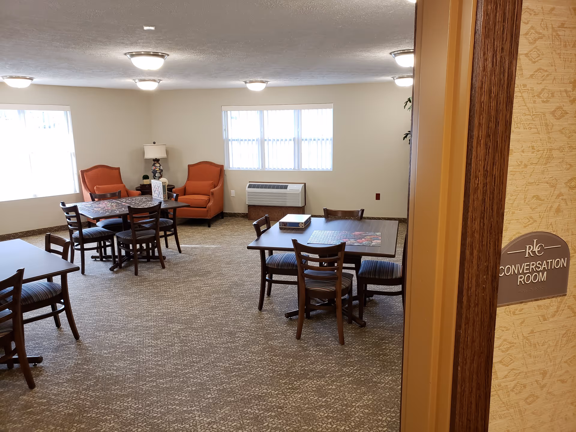 Interior view of a conversation room with several tables and chairs arranged for group activities or socializing. Two orange armchairs and a lamp are positioned near the windows, which allow natural light into the room. The room has a carpeted floor and ceiling lights. A sign on the wall near the door reads 'Conversation Room'.