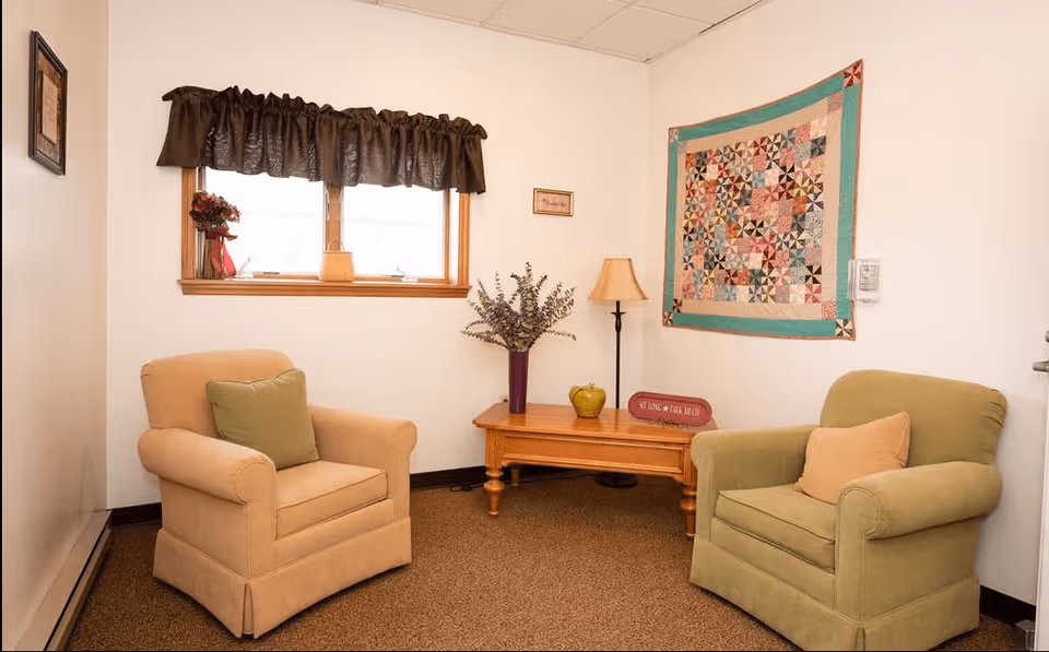 A cozy sitting area with two upholstered armchairs, one beige with a green pillow and one green with a beige pillow, positioned around a wooden coffee table. The table holds a vase with flowers, a decorative apple, a lamp, and a small sign. A window with a dark valance is on the wall behind one chair, and a colorful quilt hangs on the adjacent wall.