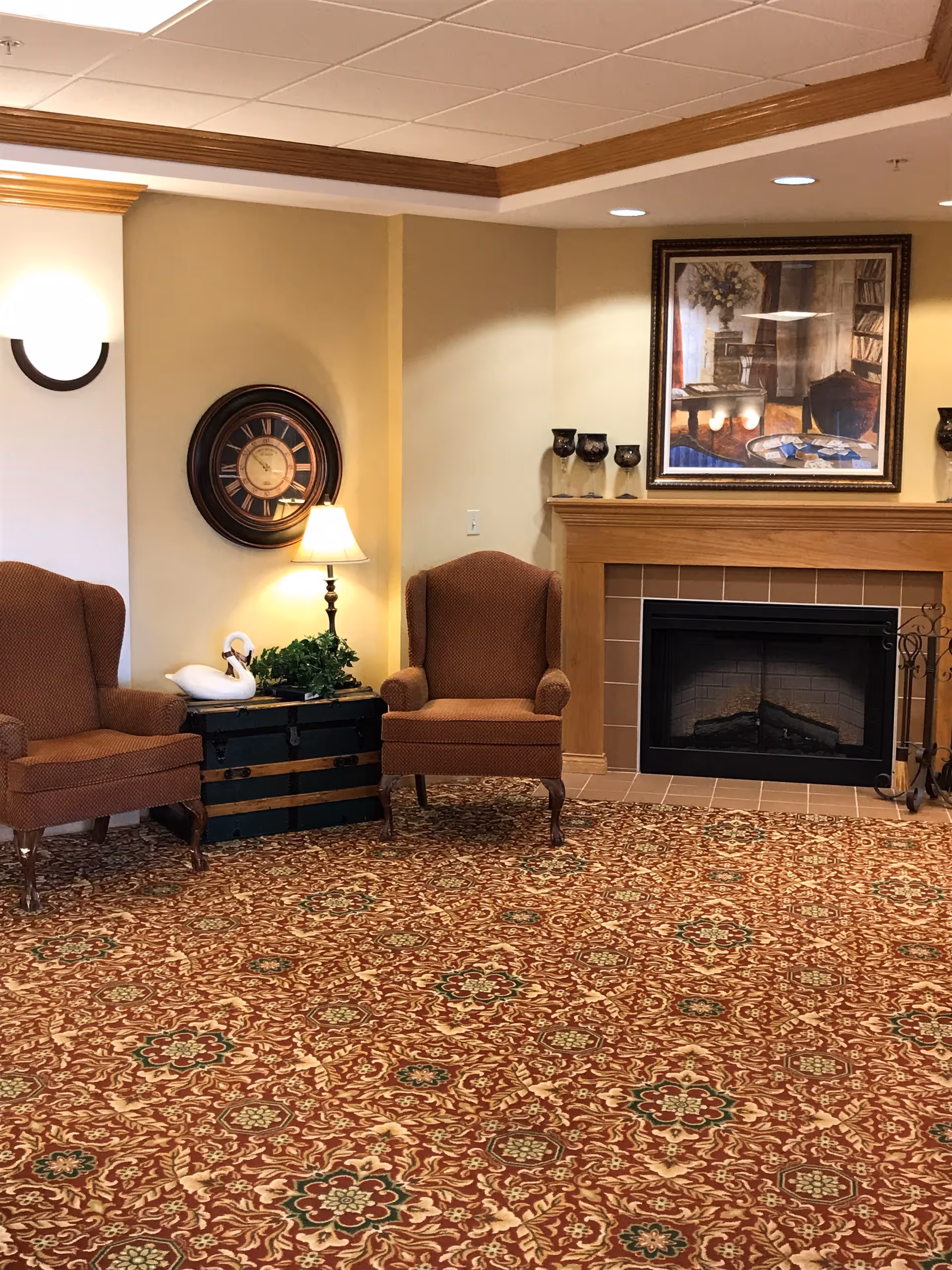 A cozy sitting area with two upholstered armchairs placed on a patterned carpet. Between the chairs is a small black chest with a table lamp, a decorative swan, and a plant. A round wall clock hangs above the lamp. To the right is a fireplace with a wooden mantle, decorated with three candle holders and a framed painting above it.