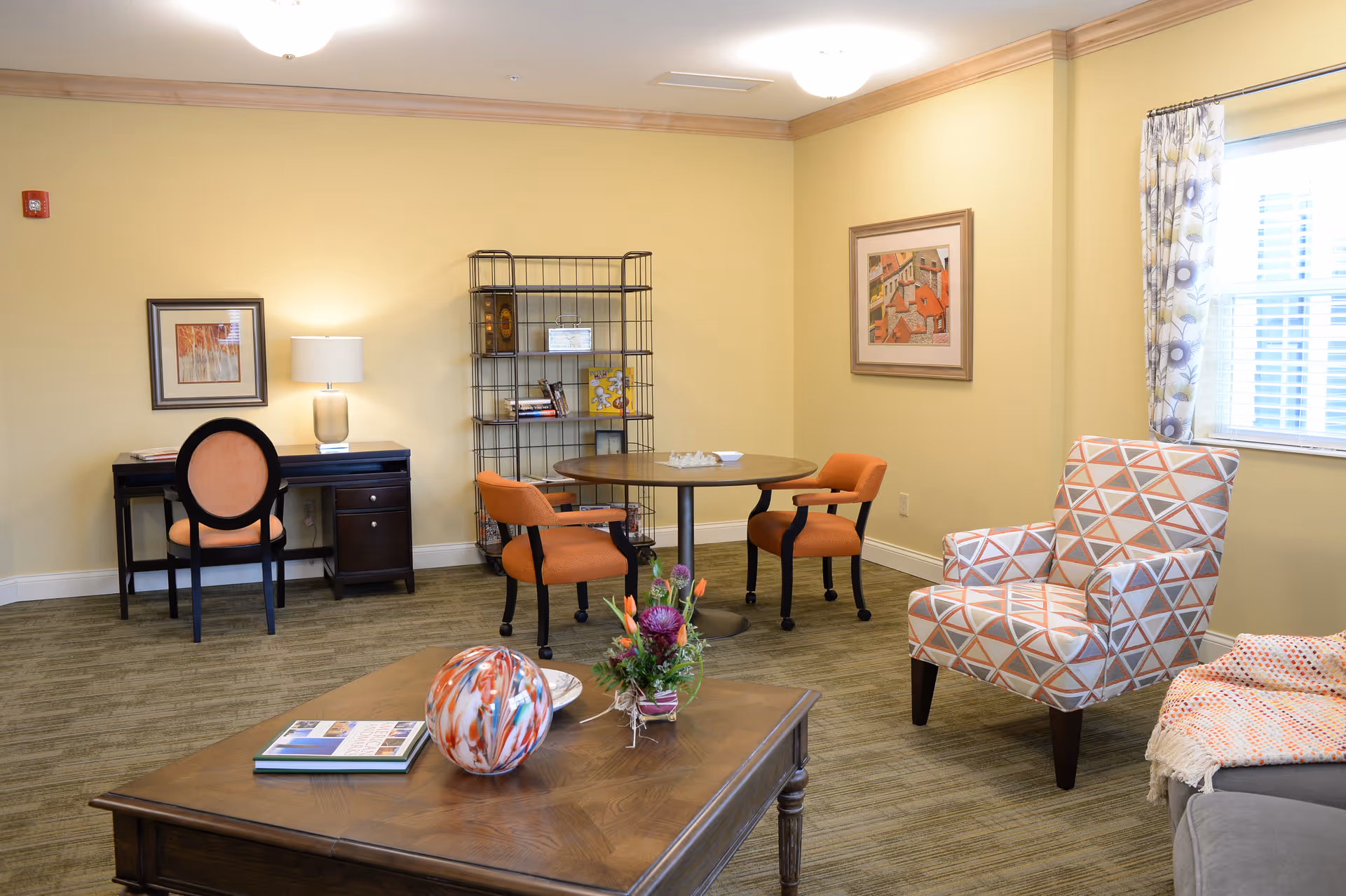 A cozy interior room with beige walls and carpeted floor featuring a wooden coffee table with a decorative glass orb and a flower arrangement. There is a patterned armchair with orange, white, and gray triangles near a window with floral curtains. In the background, there is a round table with two orange chairs, a black metal shelving unit with books and decor, and a dark wooden desk with an orange cushioned chair and a table lamp. Two framed artworks hang on the walls.